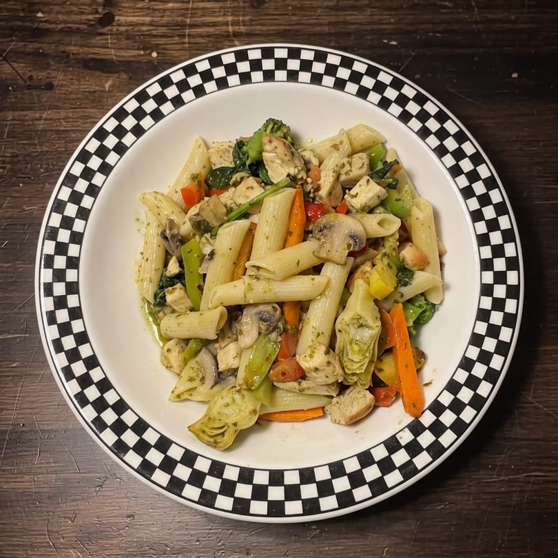 A bowl of penne pasta with chicken, broccoli, carrots, mushrooms, and artichokes in pesto sauce.
