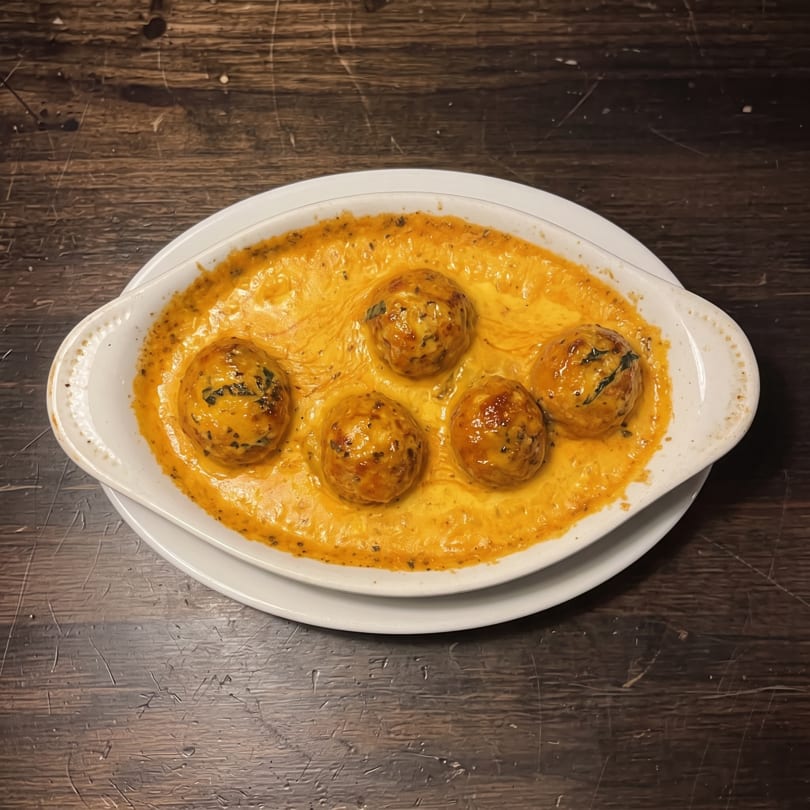 Five golden-brown baked balls in a rich, creamy orange sauce, presented in a white oval dish.
