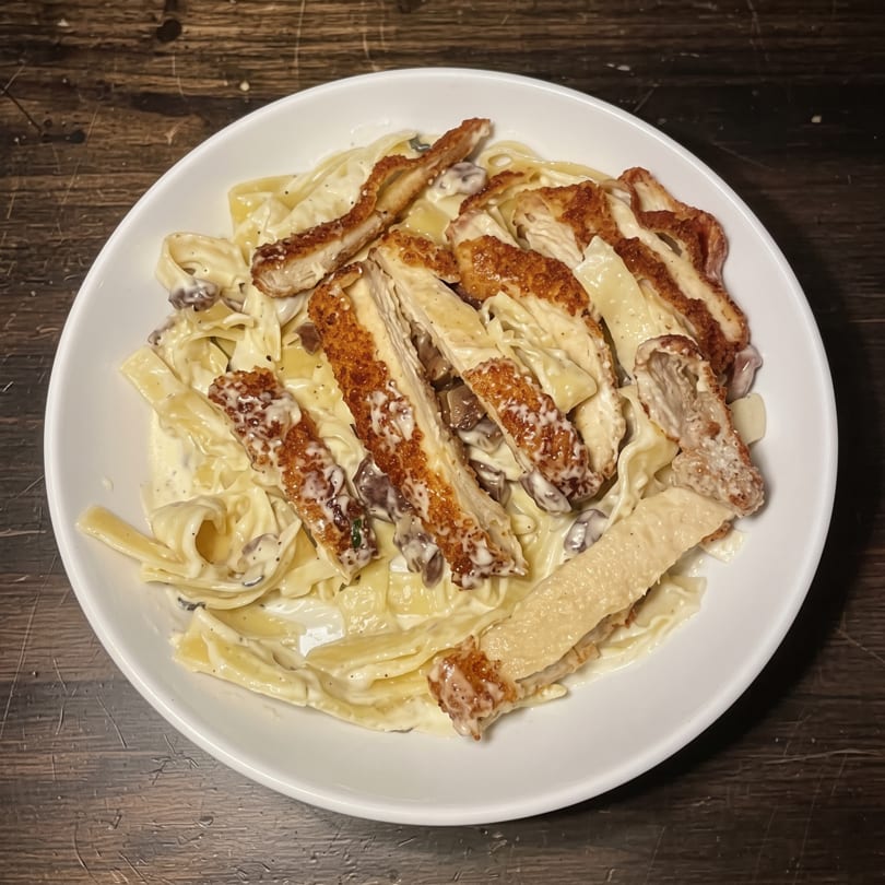 Creamy fettuccine pasta with sliced breaded chicken and mushrooms in a white bowl.